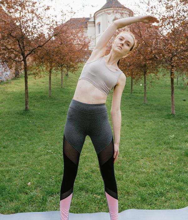 Woman doing a gentle seated stretch on a yoga mat with beautiful green plants in the background.